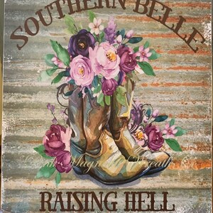 May include: A rustic wooden sign with the text "Southern Belle Raising Hell" in brown lettering. The sign features a pair of brown cowboy boots with a bouquet of pink, purple, and white flowers.