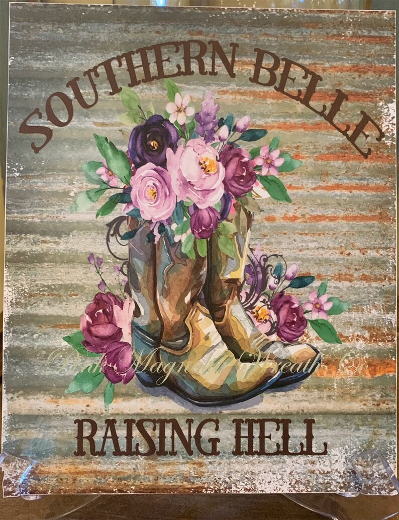 May include: A rustic wooden sign with the text "Southern Belle Raising Hell" in brown lettering. The sign features a pair of brown cowboy boots with a bouquet of pink, purple, and white flowers.