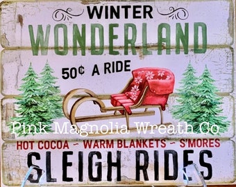May include: A rustic wooden sign with the words "WINTER WONDERLAND" in green. It features a red sled with a floral design, two green Christmas trees, and the text "SLEIGH RIDES". The sign also includes the text "HOT COCOA - WARM BLANKETS - S'MORES".