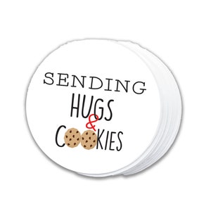 Pode incluir: Etiqueta redonda branca com texto preto que diz "Sending Hugs & Cookies" com dois biscoitos de chocolate.