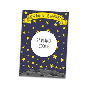 Puede incluir: Una tarjeta azul con estrellas amarillas y una pancarta amarilla que dice "Best Dad in the Universe". La tarjeta tiene un círculo blanco con el texto "2" Planet Cookie" en el centro. La parte inferior de la tarjeta tiene una luna gris con cráteres.