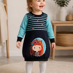 Könnte beinhalten: Ein Kleinkind trägt ein marineblaues und türkisfarbenes Kleid mit einer Cartoon-Illustration eines Mädchens mit Blumenkranz. Das Kleid hat türkisfarbene Ärmel und ein gestreiftes Oberteil. Das Kind trägt außerdem weiße Leggings und weiße Turnschuhe.