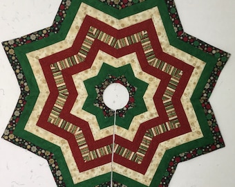 May include: A star-shaped quilted Christmas tree skirt with a green, red, and cream color scheme. The skirt has a snowflake pattern and a hole in the center for the tree trunk.