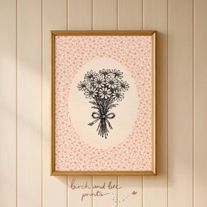 May include: A framed print featuring a black and white illustration of a daisy bouquet tied with a bow. The background is a pink floral pattern. The print is in a gold frame. The text "birch and bee prints" is at the bottom.