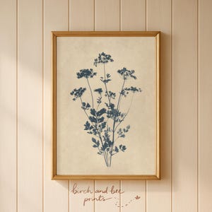 Puede incluir: Una l&aacute;mina de arte mural enmarcada que presenta una ilustraci&oacute;n bot&aacute;nica en azul oscuro de un grupo de flores silvestres sobre un fondo beige claro texturizado. La obra est&aacute; en un marco fino de metal dorado y muestra una silueta vegetal detallada.