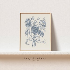 Puede incluir: Una impresi&oacute;n floral vintage azul y blanca con un ramo de rosas atado con una cinta. La impresi&oacute;n est&aacute; enmarcada en un marco de madera claro.