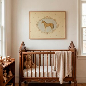 May include: A rectangular wall art print in a light wood frame featuring a tan horse illustration centered within an ornate floral border on a cream background with small repeating floral motifs, displayed above a wooden crib with a beige blanket.