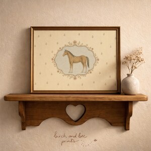 May include: A framed print featuring a brown horse illustration inside an ornate floral oval border on a cream background with small flower motifs. The print is displayed on a wooden shelf with a heart-shaped cutout, next to a small ceramic vase.