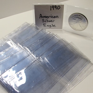 K&ouml;nnte beinhalten: Eine Sammlung klarer Kunststoff-M&uuml;nzhalter, mit einer 1990 American Silver Eagle-M&uuml;nze in einem. Ein handgeschriebenes Schild zeigt "1990 American Silver Eagle". Die Halter liegen auf einer wei&szlig;en Oberfl&auml;che.
