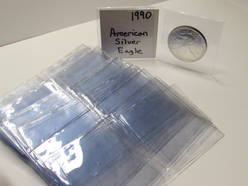 K&ouml;nnte beinhalten: Eine Sammlung klarer Kunststoff-M&uuml;nzhalter, mit einer 1990 American Silver Eagle-M&uuml;nze in einem. Ein handgeschriebenes Schild zeigt "1990 American Silver Eagle". Die Halter liegen auf einer wei&szlig;en Oberfl&auml;che.