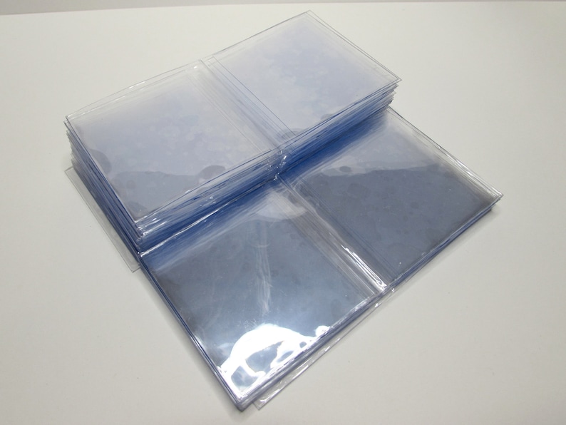 K&ouml;nnte beinhalten: Ein Stapel klarer, quadratischer Kunststoffh&uuml;llen, wahrscheinlich zur Aufbewahrung von Karten oder Fotos. Die H&uuml;llen sind transparent und erm&ouml;glichen die Sichtbarkeit des Inhalts. Das Bild ist aus leicht erh&ouml;hter Perspektive aufgenommen und zeigt den Stapel und einige einzelne H&uuml;llen.