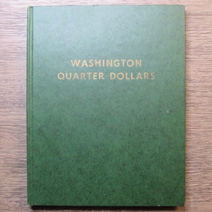 May include: A green book with the words "WASHINGTON QUARTER DOLLARS" in gold lettering. The book is closed and has a textured surface. The book is likely a collector's album for Washington quarter dollars.