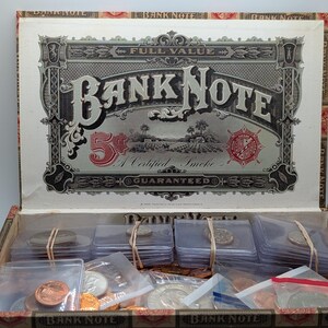 Könnte beinhalten: Geöffnete antike Bank Note-Box, gefüllt mit verschiedenen Münzen in Schutzhüllen. Der Deckel der Box zeigt verzierte Texte und Grafiken, darunter "Bank Note" und "5¢". Die Box besteht aus Pappe mit einem dekorativen Rand.