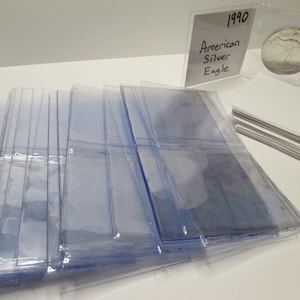 K&ouml;nnte beinhalten: Eine Ansammlung klarer Plastikh&uuml;llen, wahrscheinlich zur Aufbewahrung von Karten oder Sammlerst&uuml;cken, liegt auf einer wei&szlig;en Oberfl&auml;che. Eine Silberm&uuml;nze in einer Schutzh&uuml;lle und ein Stapel wei&szlig;er Karten sind ebenfalls zu sehen. Ein handgeschriebenes Schild tr&auml;gt die Aufschrift "1990 American Silver Eagle."