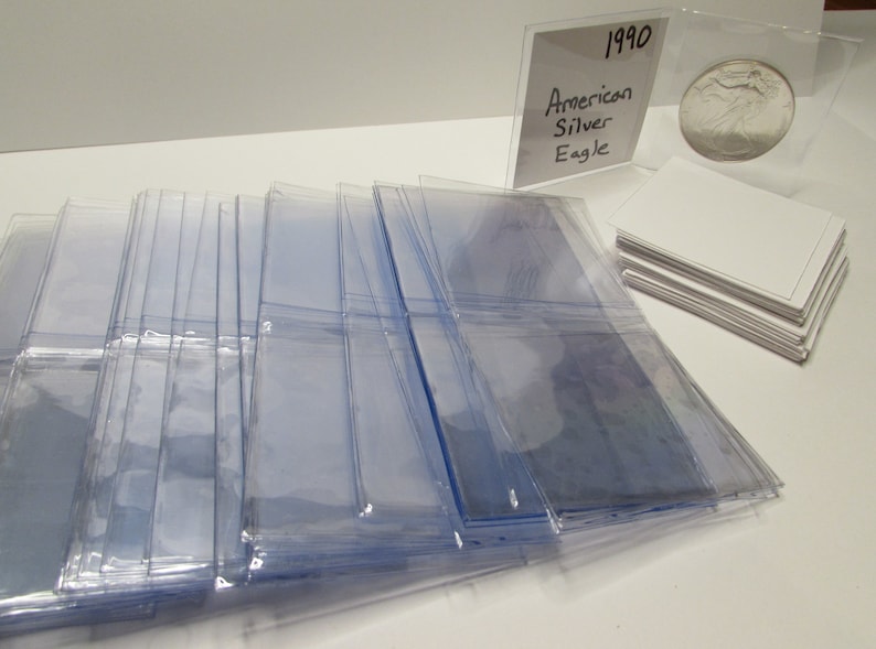 K&ouml;nnte beinhalten: Eine Ansammlung klarer Plastikh&uuml;llen, wahrscheinlich zur Aufbewahrung von Karten oder Sammlerst&uuml;cken, liegt auf einer wei&szlig;en Oberfl&auml;che. Eine Silberm&uuml;nze in einer Schutzh&uuml;lle und ein Stapel wei&szlig;er Karten sind ebenfalls zu sehen. Ein handgeschriebenes Schild tr&auml;gt die Aufschrift "1990 American Silver Eagle."