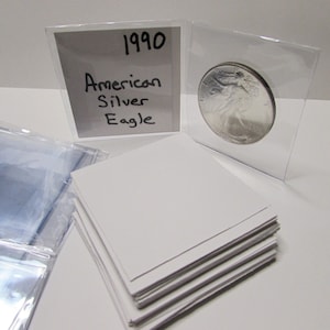 K&ouml;nnte beinhalten: Eine silberne American Eagle M&uuml;nze von 1990 in einer Schutzh&uuml;lle. Die M&uuml;nze wird mit einer wei&szlig;en Karte mit der Aufschrift "American Silver Eagle" und dem Jahr "1990" pr&auml;sentiert. Zus&auml;tzliche transparente H&uuml;llen und wei&szlig;e Karten sind ebenfalls sichtbar.