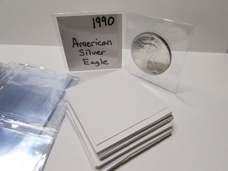 K&ouml;nnte beinhalten: Eine silberne American Eagle M&uuml;nze von 1990 in einer Schutzh&uuml;lle. Die M&uuml;nze wird mit einer wei&szlig;en Karte mit der Aufschrift "American Silver Eagle" und dem Jahr "1990" pr&auml;sentiert. Zus&auml;tzliche transparente H&uuml;llen und wei&szlig;e Karten sind ebenfalls sichtbar.