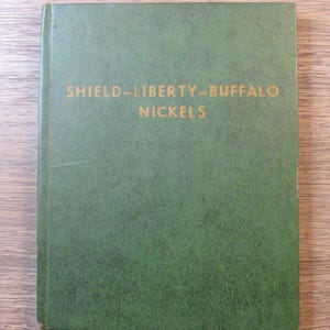 Puede incluir: Un libro verde con letras doradas que dice "SHIELD-LIBERTY-BUFFALO NICKELS". El libro tiene una cubierta texturizada, probablemente un álbum o guía de coleccionismo de monedas.