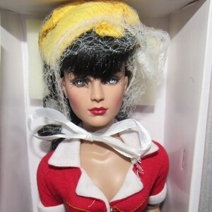 May include: A Wonder Woman doll in a display box. The doll wears a red top with white trim, a gold belt, and a blue skirt. She has black hair, blue eyes, and a yellow hat with a veil. A white ribbon is tied around her neck.