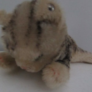 Pode incluir: Um pequeno filhote de tigre de pelúcia com um padrão de listras marrom claro e cinza. O brinquedo tem um olho de botão e está deitado de lado com a cabeça apoiada nas patas.
