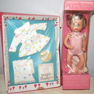 May include: Vintage Teenie Weenie Tiny Tears doll and accessories. The doll is in a pink box and wears a pink onesie. The accessories include a white robe and towel with colourful polka dots, and a "Magic Shampoo" label.