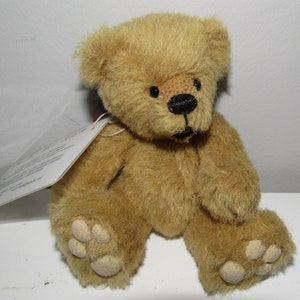 May include: A small, brown teddy bear with black eyes and a black nose. The bear is sitting on a white surface.