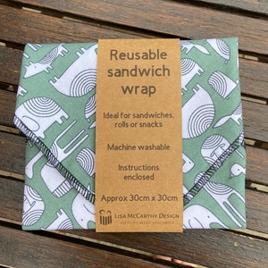 Può includere: Avvolgitore per sandwich riutilizzabile con un motivo di animali stilizzati verde e bianco. L'avvolgitore è lavabile in lavatrice e misura circa 30 cm x 30 cm. Il testo sull'etichetta recita "Avvolgitore per sandwich riutilizzabile", "Ideale per sandwich, panini o snack", "Lavabile in lavatrice", "Istruzioni incluse", "Circa 30 cm x 30 cm", "LISA MCCARTHY DESIGN", "GIFTS TO MAKE YOU SMILE".