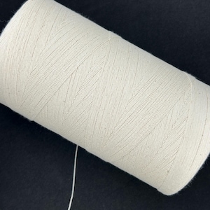 Op de afbeelding: Een close-up van een grote spoel crèmekleurig garen. Het garen is strak gewikkeld, met zichtbare diagonale lijnen. Een enkele draad loopt van de spoel. De achtergrond is effen zwart.