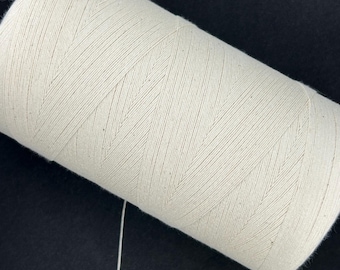 Könnte beinhalten: Nahaufnahme einer großen Spule cremefarbenen Fadens. Der Faden ist eng gewickelt, mit sichtbaren diagonalen Linien. Ein einzelner Faden erstreckt sich von der Spule. Der Hintergrund ist einfarbig schwarz.