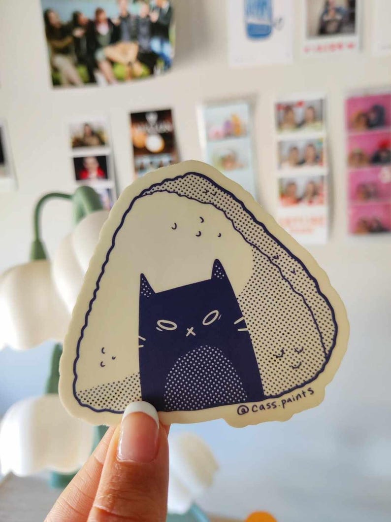 May include: A beige and white onigiri sticker with a black cat inside. The cat has a black nose and white eyes with an X in the middle. The sticker has a dotted pattern and the text "@cass.paints" at the bottom.
