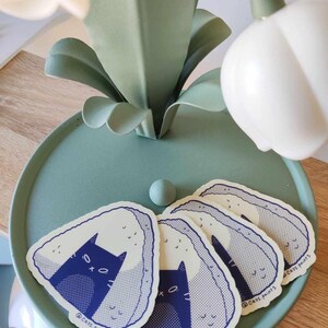 May include: Five stickers featuring a cartoon cat inside a rice ball. The rice balls are white with blue and white speckles. The cat is blue with white eyes and a white mouth. The stickers are on a light blue surface. The text on the stickers reads "@cass_paints".