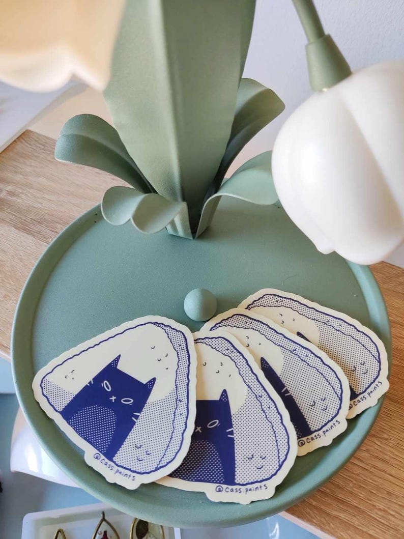 May include: Five stickers featuring a cartoon cat inside a rice ball. The rice balls are white with blue and white speckles. The cat is blue with white eyes and a white mouth. The stickers are on a light blue surface. The text on the stickers reads "@cass_paints".