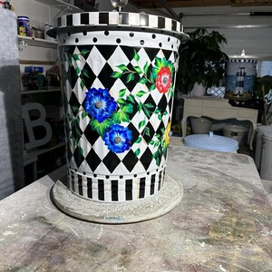 May include: A white metal trash can with a black and white diamond pattern and floral designs. The can has a black and white striped band around the bottom and a black lid with white polka dots. The can is sitting on a gray surface.
