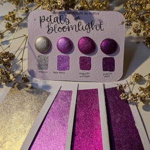 May include: A watercolour palette featuring four shimmering colours: Moonlit, Rose Petal, Magenta Whisper, and Purple Rain. The palette is labelled "petals in bloomlight" and includes colour swatches. Gold floral decorations surround the palette.
