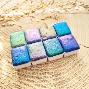 Op de afbeelding: Een set van acht iriserende aquarelverf in verschillende tinten blauw, groen en paars. De verf is in een vierkante vorm op een houten oppervlak gerangschikt.