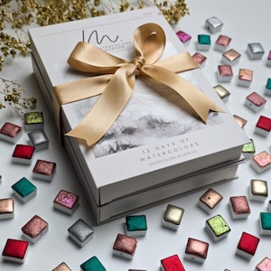 Op de afbeelding: Een witte rechthoekige doos met het opschrift 12 Days of Watercolors by Myrthe, vastgebonden met een gouden satijnen lint. De doos is omringd door talloze kleine, vierkante aquarelverf napjes in diverse metallic en levendige kleuren.