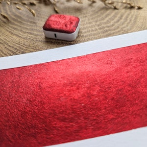 Op de afbeelding: Een kleine, vierkante, rode aquarelverfpan met een witte plastic behuizing ligt op een houten oppervlak. Daaronder een staal van rode metallic verf op wit papier. Gedroogd goudkleurig gras accentueert de achtergrond.