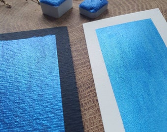 May include: Two small pans of metallic blue watercolor paint. The paint is shown on a black and white paper and a white paper. The paint is a vibrant blue with a metallic sheen.