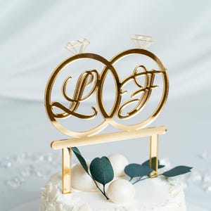 Peut inclure: Topper de gâteau de mariage en or avec deux anneaux entrelacés avec les initiales "L" et "F" et un motif de diamant. Le topper est sur un gâteau blanc avec des feuilles vertes et des boules blanches.