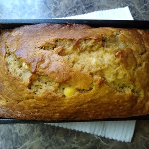 May include: A golden brown banana bread loaf baked in a black loaf pan. The bread is slightly cracked on top and has a few visible banana chunks.