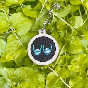 May include: A handmade keychain featuring a small, round embroidery hoop with a black fabric background. The embroidery depicts a stylized pair of breasts with blue and green accents. The hoop is made of light-colored wood and has a silver keyring.