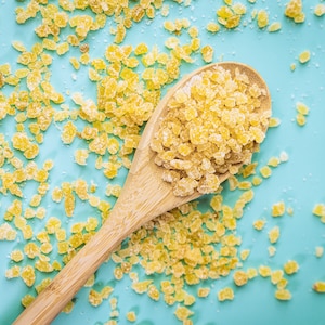 May include: A wooden spoon filled with yellow sugar crystals on a light blue background. The sugar crystals are scattered around the spoon.