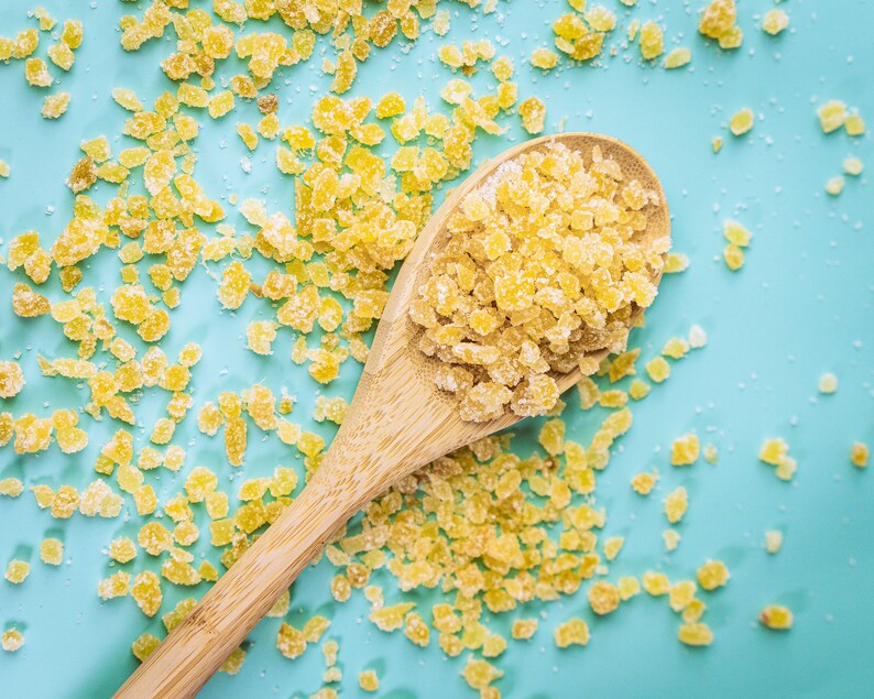 May include: A wooden spoon filled with yellow sugar crystals on a light blue background. The sugar crystals are scattered around the spoon.