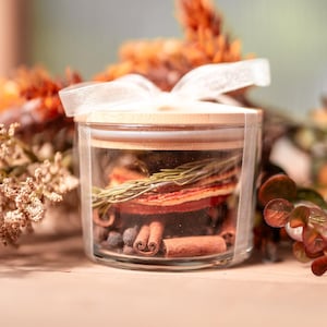 May include: A clear glass jar with a bamboo lid, tied with a white ribbon. Inside the jar are dried orange slices, a sprig of green rosemary, cinnamon sticks, and dark peppercorns, creating a layered arrangement of natural botanical ingredients.