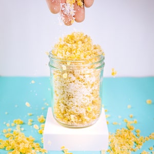 May include: A glass jar filled with yellow sugar crystals and white sugar. The jar is on a white platform and there are yellow sugar crystals scattered on a light blue surface.
