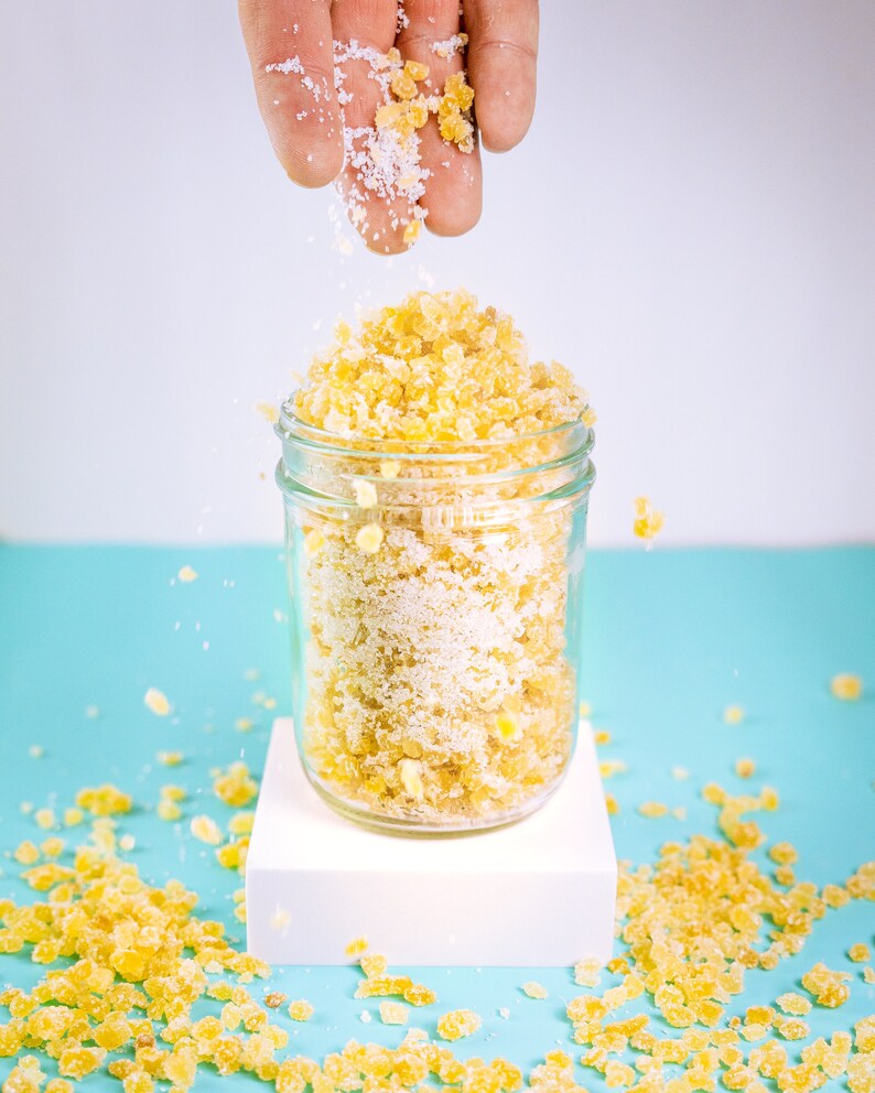 May include: A glass jar filled with yellow sugar crystals and white sugar. The jar is on a white platform and there are yellow sugar crystals scattered on a light blue surface.