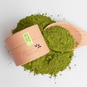 May include: A top-down view of matcha tea powder with a wooden spoon and a cylindrical container. The container is light brown with a green label that reads "Matcha". The powder is a vibrant green color, and the spoon is filled with the same powder.