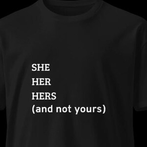 以下が含まれることがあります： 白文字で「SHE HER HERS (and not yours)」と書かれた黒いTシャツ。Tシャツは半袖でクルーネックです。テキストはシャツの前面の中央に配置されています。