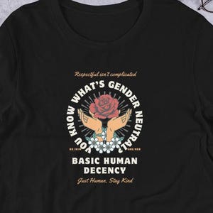 May include: Black t-shirt with a circular graphic featuring a rose, hands, and text that reads "WHAT'S GENDER NEUTRAL, BASIC HUMAN DECENCY, Just Human, Stay Kind." The shirt also has the text "Respectful isn't complicated."