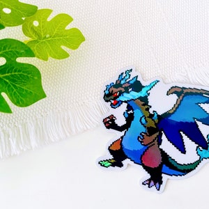 May include: A blue and brown pixelated dragon sticker with a white background. The dragon has a blue body, brown wings, and a red and yellow tail.
